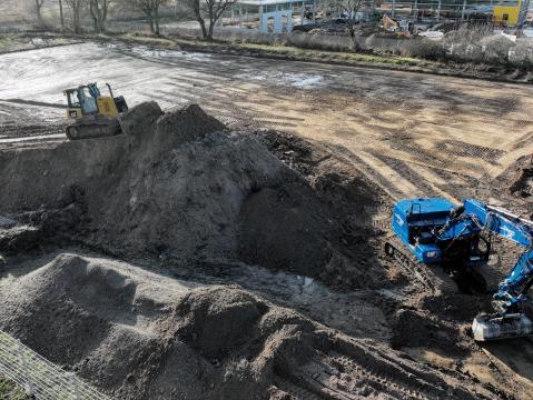 Erdarbeiten auf einer Baustelle mit einem blauen Bagger und einer gelben Planierraupe. Im Vordergrund liegen große Erdhügel, während die Baustelle von Bäumen und einem Zaun umgeben ist.