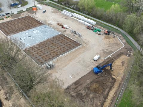 Luftaufnahme einer Baustelle mit vorbereiteten Fundamenten. Zwei große rechteckige Flächen mit Stahlarmierung dominieren das Bild. Im Hintergrund sind Baumaschinen, Baumaterialien und angrenzende Grünflächen zu sehen.