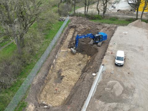 Nahaufnahme eines blauen Baggers, der in einer Baugrube arbeitet. Neben der Baustelle steht ein weißer Transporter, und im Hintergrund befindet sich ein mit Bäumen gesäumter Weg.