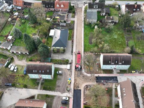 Luftaufnahme eines Wohngebiets mit mehreren Straßen und Gebäuden. Links und rechts der Straßen befinden sich Einfamilienhäuser mit Gärten sowie Reihenhäuser. In der Mitte des Bildes ist ein kleiner Parkplatz mit Autos und einem zentralen Gebäude sichtbar. Das Gebiet ist von grünen Flächen und Gärten umgeben.