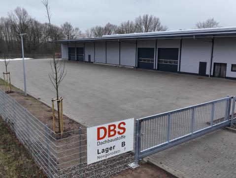 Fertige Lagerhalle mit gepflastertem Vorplatz und neu gepflanzten Bäumen entlang eines Zauns. Ein Schild mit der Aufschrift „DBS Dachbaustoffe, Lager B, Novgorodstr. 7“ steht im Vordergrund.