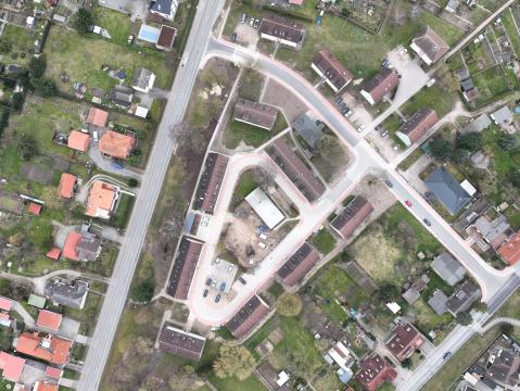Luftaufnahme eines Wohngebiets mit mehreren Straßen und Gebäuden. Links und rechts der Straßen befinden sich Einfamilienhäuser mit Gärten sowie Reihenhäuser. In der Mitte des Bildes ist ein kleiner Parkplatz mit Autos und einem zentralen Gebäude sichtbar. Das Gebiet ist von grünen Flächen und Gärten umgeben.