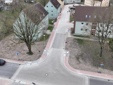 Luftaufnahme eines Wohngebiets mit mehreren Straßen und Gebäuden. Links und rechts der Straßen befinden sich Einfamilienhäuser mit Gärten sowie Reihenhäuser. In der Mitte des Bildes ist ein kleiner Parkplatz mit Autos und einem zentralen Gebäude sichtbar. Das Gebiet ist von grünen Flächen und Gärten umgeben.