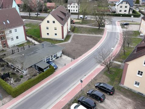 Luftaufnahme eines Wohngebiets mit mehreren Straßen und Gebäuden. Links und rechts der Straßen befinden sich Einfamilienhäuser mit Gärten sowie Reihenhäuser. In der Mitte des Bildes ist ein kleiner Parkplatz mit Autos und einem zentralen Gebäude sichtbar. Das Gebiet ist von grünen Flächen und Gärten umgeben.
