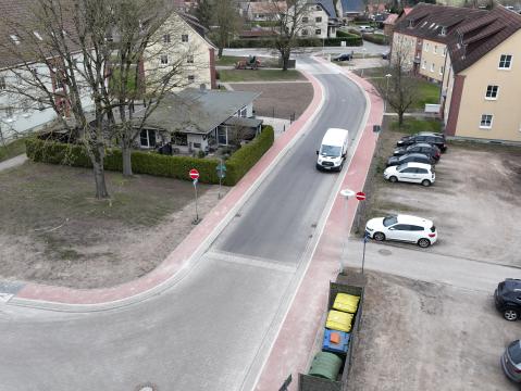 Luftaufnahme eines Wohngebiets mit mehreren Straßen und Gebäuden. Links und rechts der Straßen befinden sich Einfamilienhäuser mit Gärten sowie Reihenhäuser. In der Mitte des Bildes ist ein kleiner Parkplatz mit Autos und einem zentralen Gebäude sichtbar. Das Gebiet ist von grünen Flächen und Gärten umgeben.