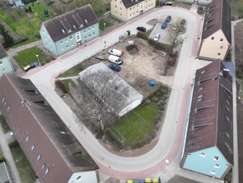 Luftaufnahme eines Wohngebiets mit mehreren Straßen und Gebäuden. Links und rechts der Straßen befinden sich Einfamilienhäuser mit Gärten sowie Reihenhäuser. In der Mitte des Bildes ist ein kleiner Parkplatz mit Autos und einem zentralen Gebäude sichtbar. Das Gebiet ist von grünen Flächen und Gärten umgeben.