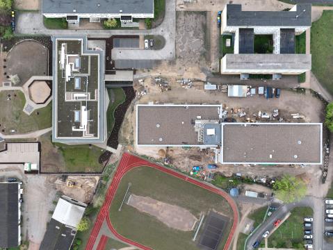 Senkrechte Luftaufnahme eines Schul- oder Verwaltungsgebäudekomplexes mit begrünten Dächern, angelegten Grünflächen und einem Sportplatz mit roter Laufbahn. Rechts unten sind Bauarbeiten zu erkennen, links kleinere Gebäude und Parkflächen.