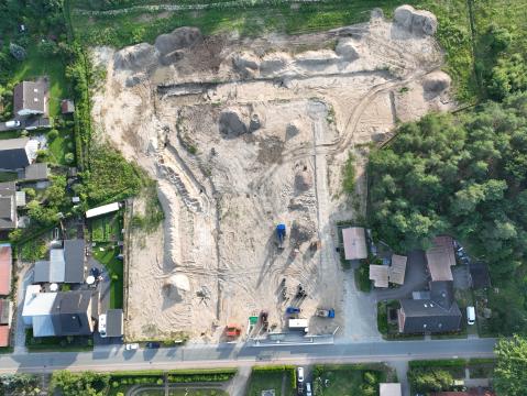  Luftaufnahme einer großen Baustelle inmitten eines Wohngebiets. Die Fläche ist gerodet und weist Spuren von Erdbewegungen auf. Umgeben wird die Baustelle von Einfamilienhäusern mit Gärten sowie angrenzendem Wald auf der rechten Seite. Baumaschinen und Materialien sind am unteren Rand der Baustelle sichtbar.