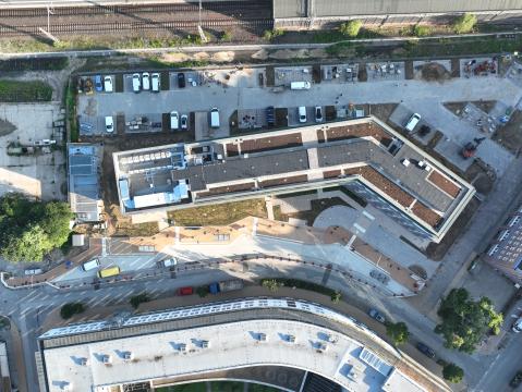 Luftaufnahme eines Bauprojekts neben einer Bahnanlage. Ein moderner Gebäudekomplex mit begrünten Dächern und einer markanten, geknickten Architektur ist im Fokus. Vor den Gebäuden befinden sich Baustellenfahrzeuge und Materialflächen, dahinter verlaufen Bahngleise.