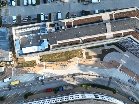 Detaillierte Luftaufnahme eines Gebäudekomplexes mit begrüntem Dach. Vor dem Gebäude ist eine neu gestaltete Fußgängerzone mit gepflasterten Wegen und Grünflächen sichtbar. Fahrzeuge und Baustellenutensilien sind auf der angrenzenden Fläche verteilt.