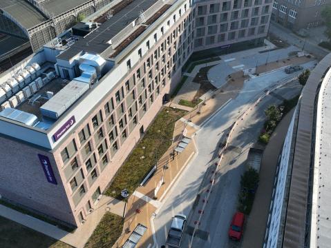 Perspektivansicht eines modernen Backsteingebäudes mit einem angrenzenden Gehweg in Bau. Im Vordergrund ist ein abgesperrter Weg mit frischen Pflastersteinen und Absperrungen sichtbar.