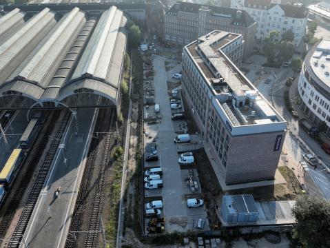 Luftaufnahme des Gebäudekomplexes in direkter Nachbarschaft eines Bahnhofs mit mehreren Bahnsteigen und Zügen. Das Hauptgebäude hat ein begrüntes Dach und ist umgeben von Parkplätzen und Baustellenbereichen.