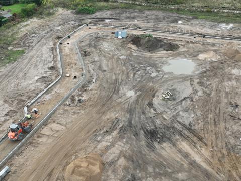 Nahaufnahme einer Baustelle, auf der Erdarbeiten für Straßen oder Wege durchgeführt werden. Im Vordergrund ist ein orangefarbener Bagger mit zwei Arbeitern zu sehen, der entlang eines frisch angelegten Bordsteins arbeitet. Mehrere Pfützen und Haufen aus Sand und Erde befinden sich auf der Baustelle.