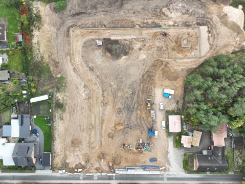 Luftaufnahme einer Baustelle in einem Wohngebiet. Zu sehen sind Erdarbeiten für ein Bauprojekt, darunter angelegte Straßengrundrisse, Baumaschinen und Materiallager. Die Baustelle grenzt an Häuser mit gepflegten Gärten und einen angrenzenden Wald.