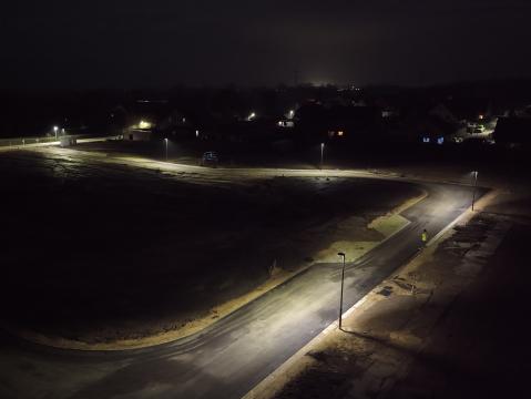 Nachtaufnahme einer neu angelegten Straße mit funktionierender Straßenbeleuchtung. Die Laternen werfen Licht auf die asphaltierte Fahrbahn, während der Rest der Umgebung dunkel bleibt. Im Hintergrund sind vereinzelt beleuchtete Häuser sichtbar.