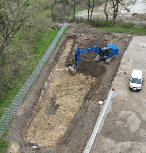 Nahaufnahme eines blauen Baggers, der in einer Baugrube arbeitet. Neben der Baustelle steht ein weißer Transporter, und im Hintergrund befindet sich ein mit Bäumen gesäumter Weg.