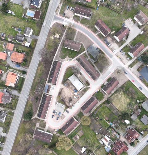 Luftaufnahme eines Wohngebiets mit mehreren Straßen und Gebäuden. Links und rechts der Straßen befinden sich Einfamilienhäuser mit Gärten sowie Reihenhäuser. In der Mitte des Bildes ist ein kleiner Parkplatz mit Autos und einem zentralen Gebäude sichtbar. Das Gebiet ist von grünen Flächen und Gärten umgeben.