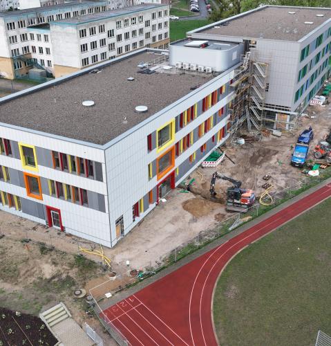 Luftaufnahme einer Baustelle mit zwei mehrstöckigen Gebäuden, deren Fassaden mit bunten Fensterrahmen in Gelb, Orange und Rot gestaltet sind. Im Vordergrund sind ein roter Sportplatz mit Laufbahn sowie Bauarbeiten mit Bagger und Baumaterialien zu sehen. Umgeben ist die Baustelle von Wohngebäuden und Parkplätzen.
