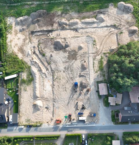  Luftaufnahme einer großen Baustelle inmitten eines Wohngebiets. Die Fläche ist gerodet und weist Spuren von Erdbewegungen auf. Umgeben wird die Baustelle von Einfamilienhäusern mit Gärten sowie angrenzendem Wald auf der rechten Seite. Baumaschinen und Materialien sind am unteren Rand der Baustelle sichtbar.