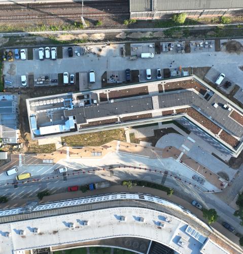 Luftaufnahme eines Bauprojekts neben einer Bahnanlage. Ein moderner Gebäudekomplex mit begrünten Dächern und einer markanten, geknickten Architektur ist im Fokus. Vor den Gebäuden befinden sich Baustellenfahrzeuge und Materialflächen, dahinter verlaufen Bahngleise.