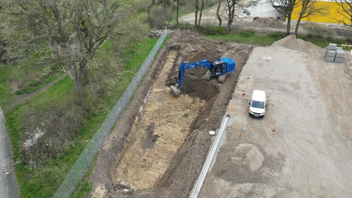 Nahaufnahme eines blauen Baggers, der in einer Baugrube arbeitet. Neben der Baustelle steht ein weißer Transporter, und im Hintergrund befindet sich ein mit Bäumen gesäumter Weg.