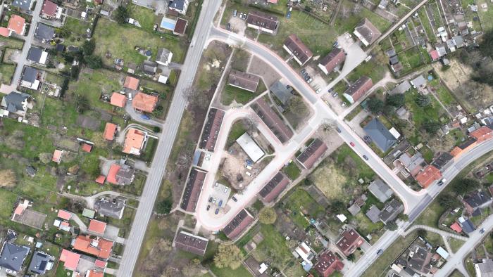 Luftaufnahme eines Wohngebiets mit mehreren Straßen und Gebäuden. Links und rechts der Straßen befinden sich Einfamilienhäuser mit Gärten sowie Reihenhäuser. In der Mitte des Bildes ist ein kleiner Parkplatz mit Autos und einem zentralen Gebäude sichtbar. Das Gebiet ist von grünen Flächen und Gärten umgeben.