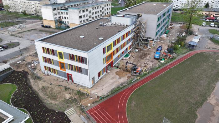 Luftaufnahme einer Baustelle mit zwei mehrstöckigen Gebäuden, deren Fassaden mit bunten Fensterrahmen in Gelb, Orange und Rot gestaltet sind. Im Vordergrund sind ein roter Sportplatz mit Laufbahn sowie Bauarbeiten mit Bagger und Baumaterialien zu sehen. Umgeben ist die Baustelle von Wohngebäuden und Parkplätzen.