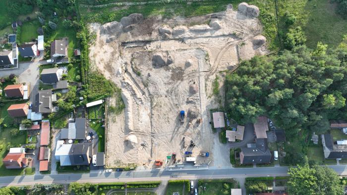  Luftaufnahme einer großen Baustelle inmitten eines Wohngebiets. Die Fläche ist gerodet und weist Spuren von Erdbewegungen auf. Umgeben wird die Baustelle von Einfamilienhäusern mit Gärten sowie angrenzendem Wald auf der rechten Seite. Baumaschinen und Materialien sind am unteren Rand der Baustelle sichtbar.