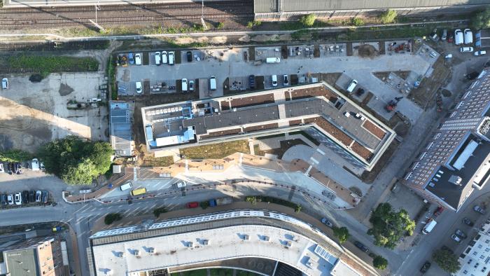 Luftaufnahme eines Bauprojekts neben einer Bahnanlage. Ein moderner Gebäudekomplex mit begrünten Dächern und einer markanten, geknickten Architektur ist im Fokus. Vor den Gebäuden befinden sich Baustellenfahrzeuge und Materialflächen, dahinter verlaufen Bahngleise.